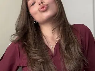 Young woman with long brown hair wearing a maroon blouse and silver necklace making a playful kissy face.