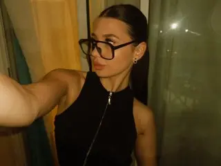 Anasteysha21 poses in a black sleeveless top, looking at the camera with her dark hair down in a dimly lit room.