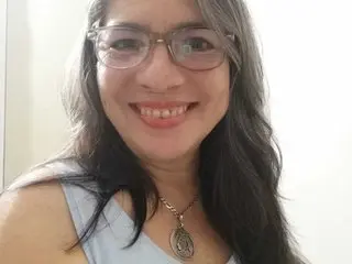AndreaLoveLatin smiles with long dark hair and glasses, wearing a light blue tank top and a silver pendant necklace.