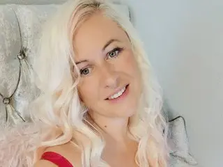 Angel34gnatural smiles with her long blonde hair, wearing a red top against a soft grey background.