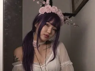 AngelAsia poses with long purple hair, wearing a white lace top and a white choker, looking directly at the camera.