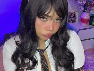 AngelDusstt poses with long dark hair and bangs, wearing a white button-down shirt with a dark tie, looking directly at the camera.