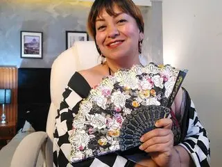 AngelVelvet smiles while holding an open decorative fan up to her chest, wearing a black and white checkered top with her brown hair styled down.