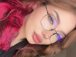 Young woman with pink hair and round glasses smiling in a soft, aesthetic close-up portrait.