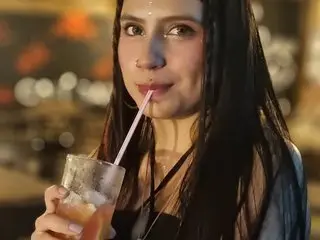 AngelaMillerx looks at the camera while holding a drink with a straw, showing her long dark hair and bare shoulders in a dimly lit setting.