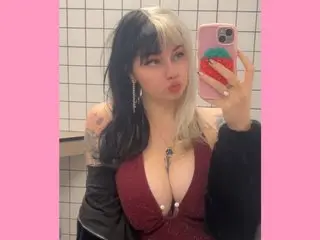 Angelina_Glass poses in a low-cut red top, showing cleavage and her long dark hair with blonde streaks, while holding a phone in a tiled bathroom.