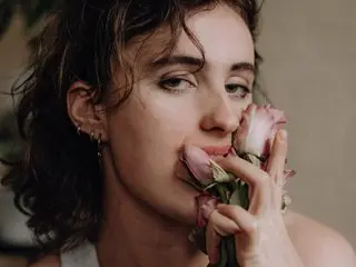 AngellLace poses with her hand near her mouth, showing her dark wavy hair and clear skin in a close-up shot.