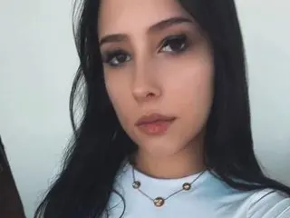 Young woman with long dark hair wearing a white top and gold necklace against a plain white background.