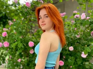 Angelsssyess with long red hair poses in a light blue tank top against a background of pink roses.