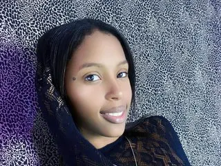 Angeya_Petite smiles at the camera with her dark hair pulled back, wearing a black lace top against a textured grey background.