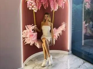 AnnSoleil poses in a white lace teddy, sitting on a pink velvet chair against a pink floral backdrop.