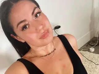 AnnaGloss poses close-up, looking at the camera with her dark hair down and wearing a black top against a plain white wall.