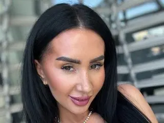 Smiling woman with long black hair and glamorous makeup posing outdoors in a modern urban setting.