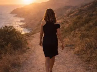 AnnabelleWarick walks away from the camera on a dirt path overlooking a coastal cliffside at sunset, wearing a short black sleeveless dress.