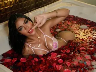 AnnieMilan poses naked in a white bathtub filled with red rose petals, wearing a pink lace bra and a delicate necklace.