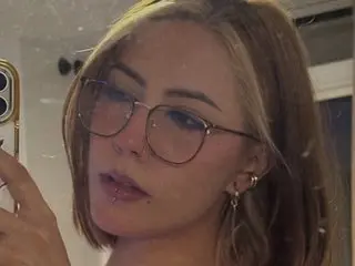 Annie_Oz poses with a direct gaze, showing her face and blonde hair while wearing thin-rimmed glasses in a close-up shot.