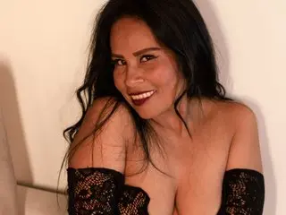 Smiling woman in a black lace off-the-shoulder outfit posing against a white wall in soft indoor lighting.