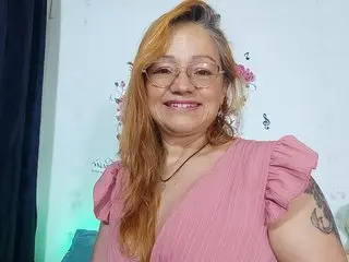 Smiling woman with blonde hair and glasses wearing a pink ruffled blouse in a bright indoor setting.