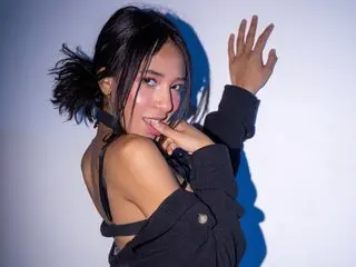AntonellaBaronee poses with her hand near her mouth, wearing a black top against a plain background.