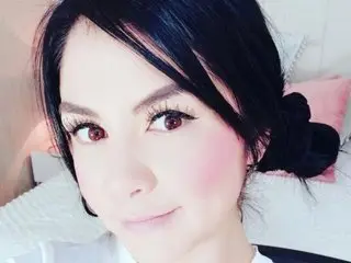 AntonellaFerreira poses close-up with her dark hair pulled back, showing her face and shoulders against a soft pink background.