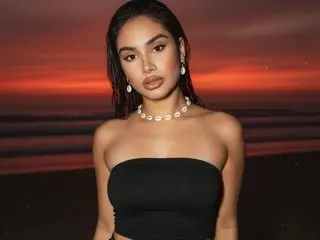 Young woman in a black tube top and shell necklace posing against a dramatic sunset beach background.