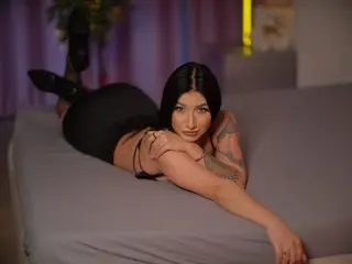 Tattooed woman in black bodysuit lounging on a grey bed in a moody, dimly lit room.