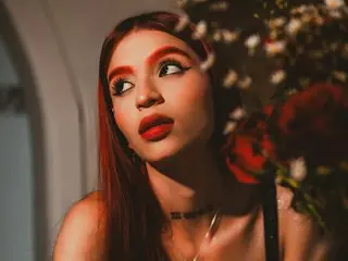 Apriiljonnes with long red hair and bold makeup poses near a bouquet of flowers.