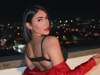 AriaMatheuss poses with her dark hair down, wearing a black bra and a red jacket against a dark night city background.
