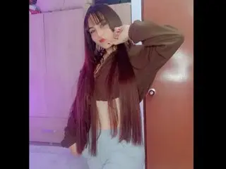 Aria_Noir poses with her hands near her face, wearing a brown long-sleeve crop top and light-colored bottoms with her long dark hair down.