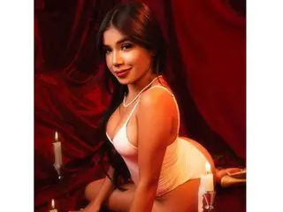 ArianaWoods poses in a white bodysuit against red curtains, smiling while sitting near lit candles.