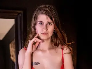 ArielXFrost poses with her hands near her face, showing her long brown hair and bare chest against a dark background.