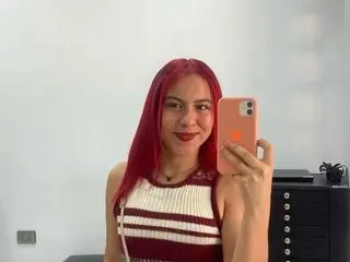 Smiling woman with vibrant red hair wearing a striped sleeveless top taking a selfie in a bright room.