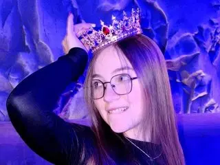 ArrinaWairr smiles while wearing glasses and a gold crown, with her long brown hair down against a blue background.