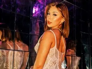 AruraSkyBabestation poses in a white lace bodysuit, showing her bare shoulders and cleavage against a dark, lit background.