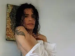 Ary_Ambrosia poses with her wet, dark hair and a white shirt pulled down to expose her shoulder and chest, showing a tattoo on her upper arm.