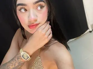 AryyySweet looks into the camera with a soft expression, wearing a gold chain top and a gold watch.