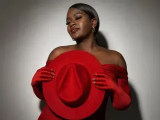 Elegant Black woman in a red off-the-shoulder dress and gloves holding a large red hat against a gray background.