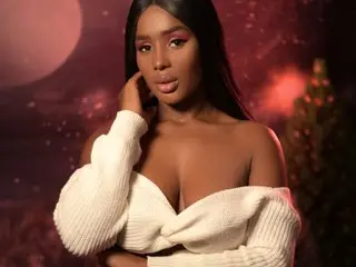 AshantiPrince23 poses with long dark hair, wearing a white off-the-shoulder knit sweater against a dark, warm-toned background.