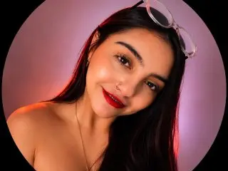 AshleeyRobertss poses with a smile, showing her long dark hair and red lipstick against a soft pink background.