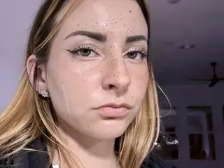 Close-up portrait of a freckled woman with winged eyeliner and blonde hair in a soft indoor setting.