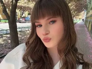 Young woman with brown bangs and wavy hair wearing a white outfit in a sunny outdoor park setting.