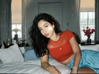 AshleyPusset poses on a bed with long dark hair, wearing a red short-sleeve top and blue shorts in a bright room.