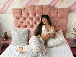 Ashly_Rousse poses on a bed wearing a white lace bra and matching lace panties, leaning back against a pink tufted headboard.