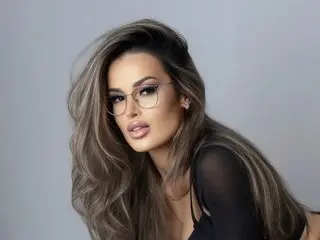 Glamorous woman with long ash-blonde hair and glasses wearing a black top against a neutral studio background.