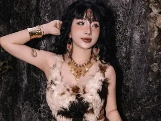 Asiangirl6969 poses with her hands near her head, wearing a white feathered top and gold jewelry against a dark, textured background.