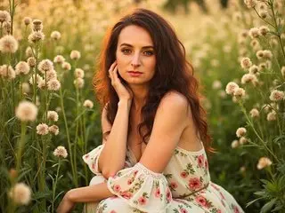 AskMeStandUp poses with her hand to her face, showing her long brown hair and white floral dress in a soft outdoor setting.