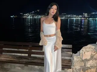 AudreyMell poses outdoors at night wearing a white crop top and matching white skirt, with her long dark hair down.