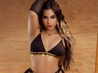 AuraSousa poses with one arm raised, wearing a black bikini top and showing her long, wavy dark hair against a plain brown background.