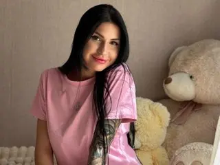 AuraXoney poses with long black hair, wearing a pink t-shirt while sitting next to a large white teddy bear.