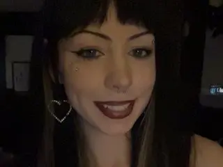 Aurorasky222 poses with a smile, showing her dark hair with bangs and dark lipstick against a dark background.